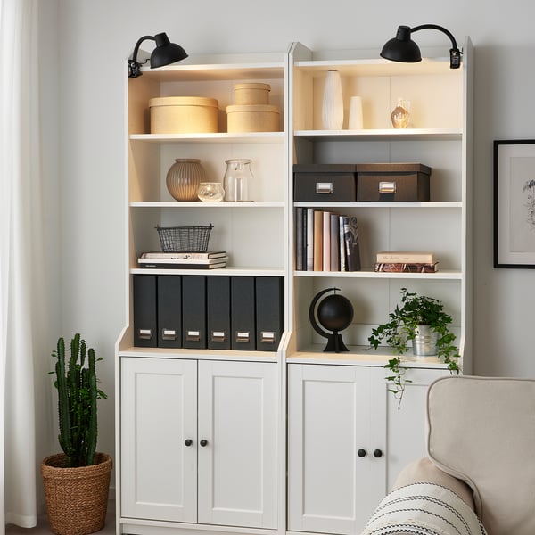 Two white SKURUP lamps illuminate a bookcase filled with books, vases, and storage boxes. A beige armchair sits beside it.