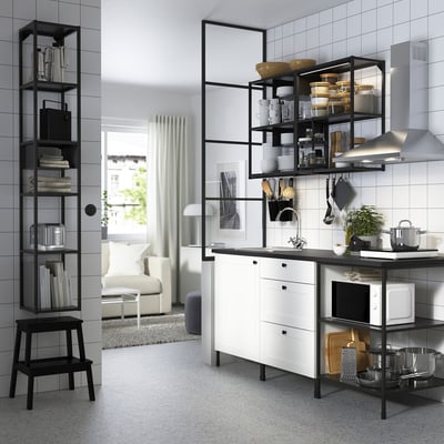 Modern kitchen: white cabinets, black accents, open shelves. Black bookshelf, stool, microwave, sink, plant. Beige sofa, rug.