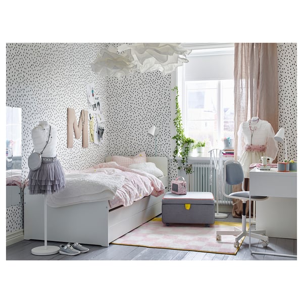 Bedroom with white bed, pink blankets, grey ottoman, desk, chair, window with curtains, plants, and wall decor.