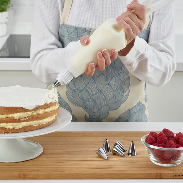 Erson decorates cake using a silicone pastry bag and metal piping tubes. Fresh raspberries nearby for topping.