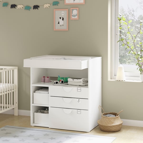 A white baby changing table with shelves and drawers. Decorated nursery with plush toys, crib, and wall art.