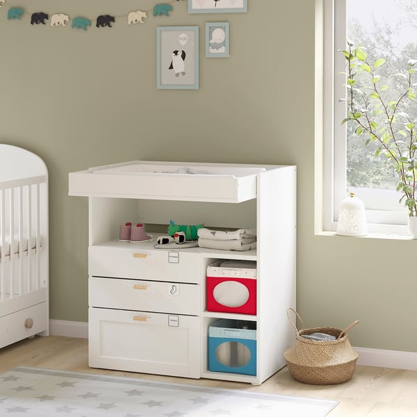 Baby room with white changing table, shelves, and toys. Green walls, wooden floor, and crib.