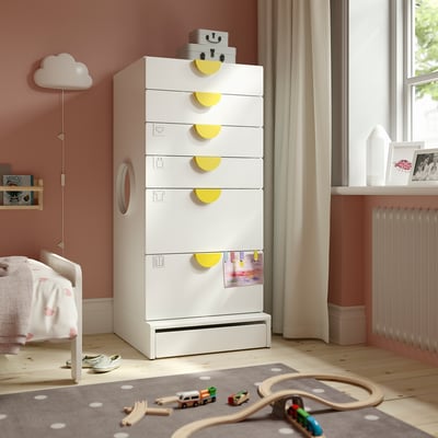 Kids room with white SMÅSTAD dresser, six drawers, yellow handles. Includes a crib, toys, books, and a window.
