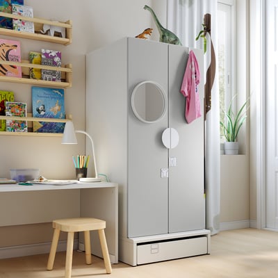 Kids room with white SMÅSTAD wardrobe, shelves filled with colourful books, small wooden stool, and desk with lamp.