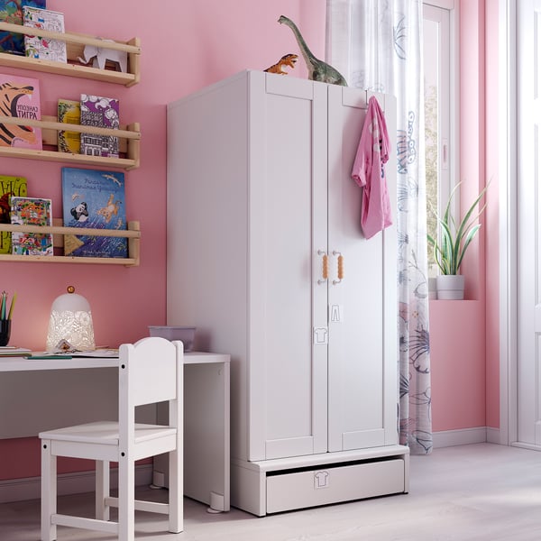 Kids room with pink walls, white SMÅSTAD wardrobe, wooden shelves, desk, and chair, dinosaur toy, plant, and curtains.