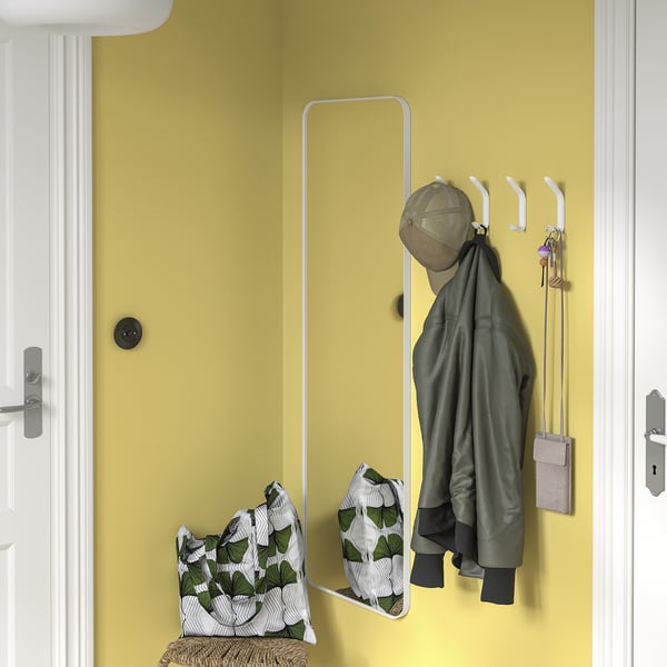 Yellow wall with white SNÖRAKA hooks holding green jacket, bag, and keys. Mirror below, wicker bench with pillow.