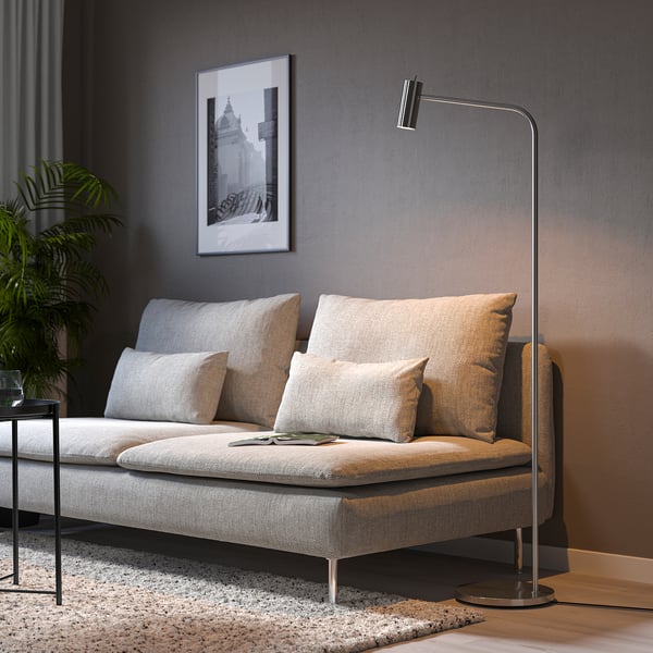 Modern grey sofa with beige cushions, black floor lamp, plant, and framed art on dark wall.