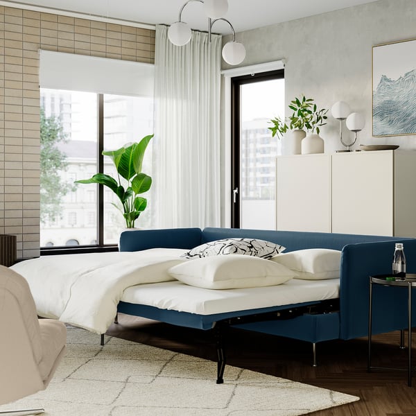 Modern bedroom with blue sofa bed, city view, plant, and stylish decor.