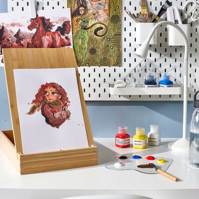 Easel with portrait, paints, brushes, canvas. Water, lamp nearby.