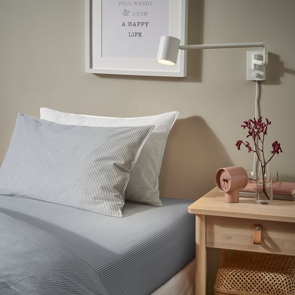Bed with striped grey pillow, wall light, vase with flowers, wooden bedside table, and wicker basket.