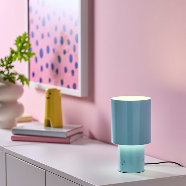 Light blue SPETSBOJ lamp sits on a white table, next to a yellow toy and books. Its small, rounded, with a dimmable shade, giving soft mood light.