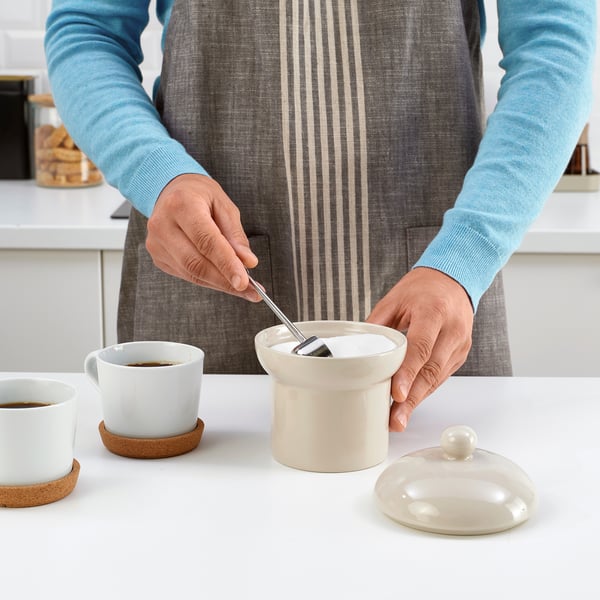 A person is using a spoon to scoop sugar from a container labelled SPINNARHAJ into a cup of coffee on a table.