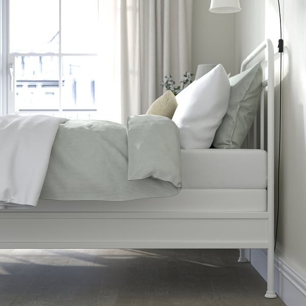 Bedroom scene featuring white metal bed frame with striped bedding, pillows, and wooden floor, beside a window with curtains.