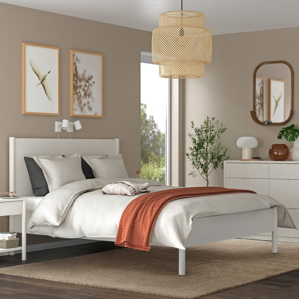 Modern bedroom with white bed, black & white pillows, red blanket, wicker light, side table lamp, dresser, nature-themed art, plant, wooden floor.
