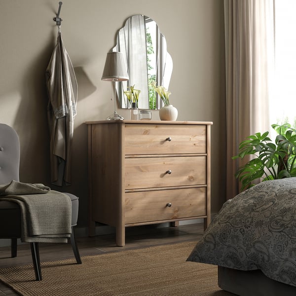 Bedroom with STOREMOLLA furniture: chest, mirror, chair, lamp, curtains, plant. Wood chest, white mirror, jute rug. Blue blanket, grey chair.