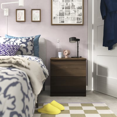 Bedroom with bed, dark wooden bedside table, lamp, clock, yellow slippers, purple walls, patterned bedding, framed photos, chequered rug.
