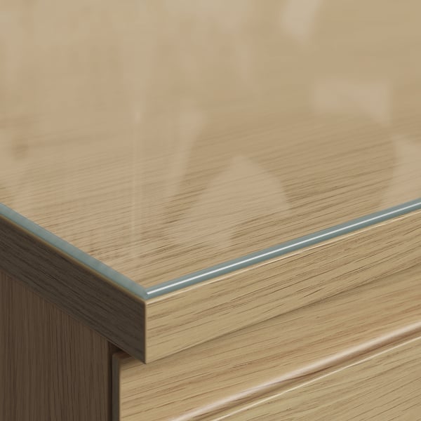 Close-up of STORKLINTA series furniture with sleek, clear glass top, showcasing light wood grain and metal edge.