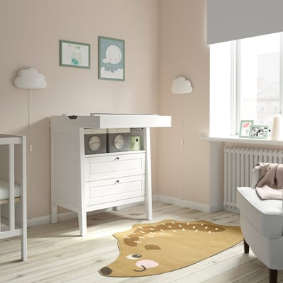 Baby nursery with SUNDVIK white chest, changing table, and deer rug. Two cloud lamps, art frames, and window with blinds. Light-colored wood floor.
