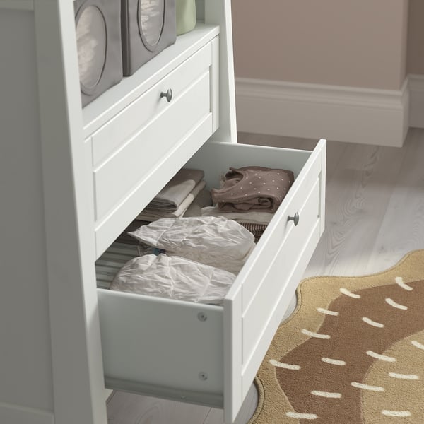 Open white drawer of baby dresser with folded clothes, on patterned rug.