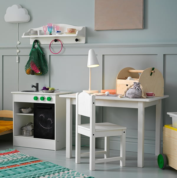 Playroom with white wooden table, chair, toy kitchen, lamp, and decorative items.