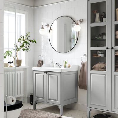 Modern bathroom with grey TÄNNFORSEN vanity, mirror, glass cabinets, plants, and windows.