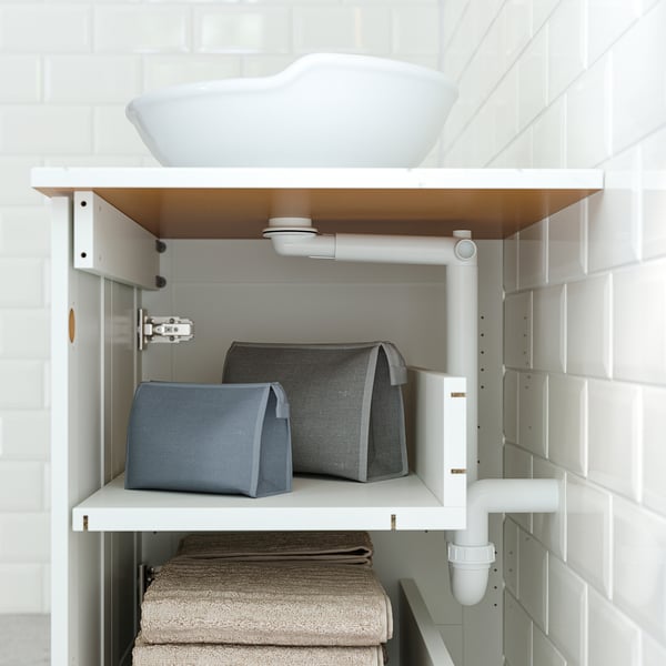 Bathroom cabinet with two shelves. Top shelf holds two fabric bins, bottom shelf stacked with towels. White pipes visible, attached to cabinet.