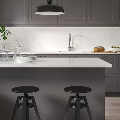 Modern kitchen island with black stools, dark cabinets, white marble, and pendant lamp. Wine glasses, plates, and bread on counter.