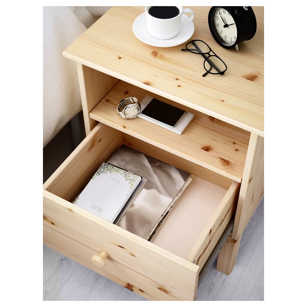 Wooden bedside table with open drawer showing book and watch; top shelf holding glasses, clock, and coffee cup.