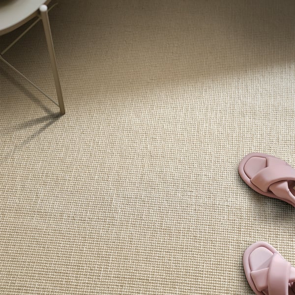 Displays a textured beige rug on a floor, with pink slippers and a chair. The rug is flatwoven, ideal for easy maintenance under furniture.