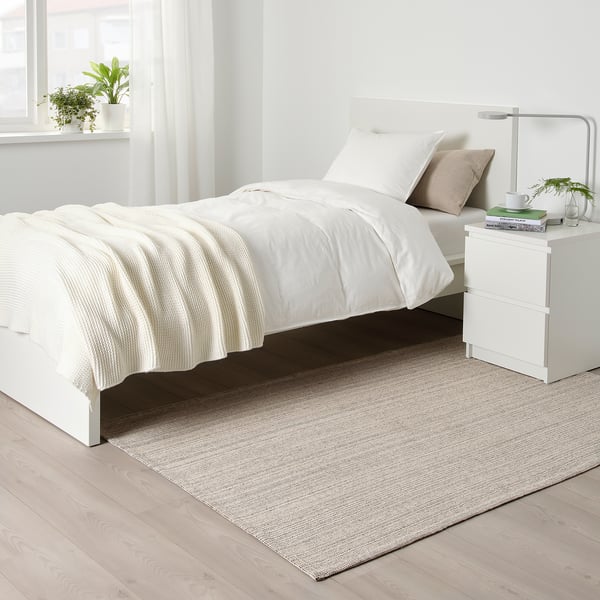 Modern minimalist bedroom with TIPHEDE rug under single bed, white sheets, beige pillow, bedside table with lamp, books, and plant.