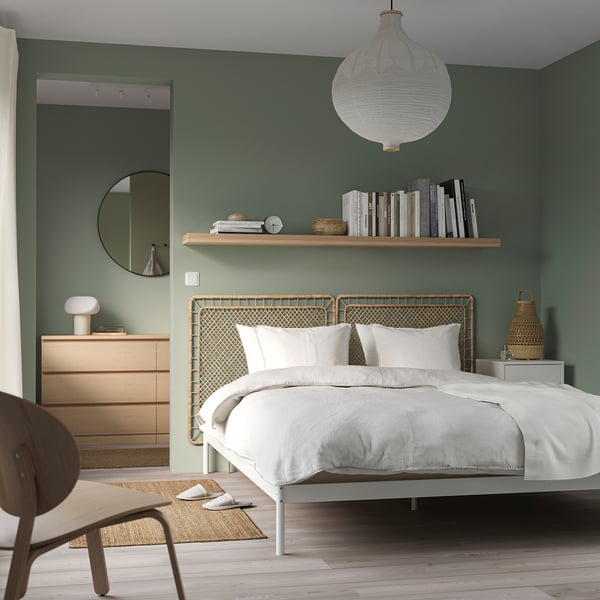 Bedroom with green walls, white bed, weaved headboard, wooden dresser, large mirror, bookshelf with books.