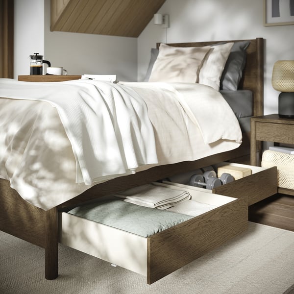 The TONSTAD bed in light brown wood, featuring two open drawers under the frame, a white bedspread, two pillows, and a wooden tray with coffee items.