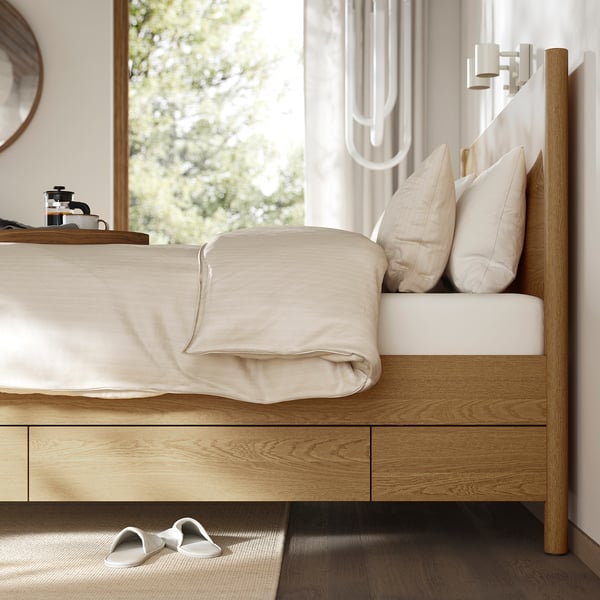 Modern wooden bed with soft-close drawers, light bedding, and slippers by window.
