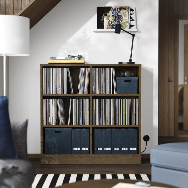A TONSTAD wooden shelf unit with vinyl records and storage boxes. Its brown, rectangular, and sits between furniture, with a lamp and chair nearby.