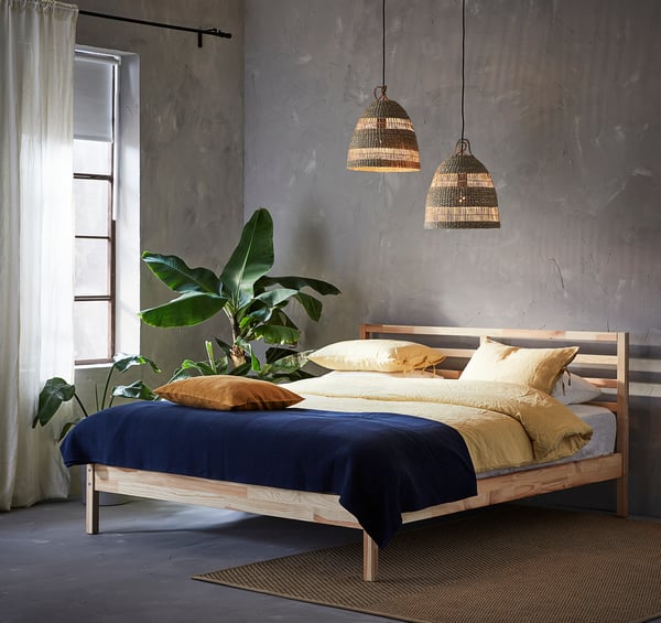 Modern bedroom with wooden bed, blue & yellow linen, hanging lamps, plant, sheer curtains.