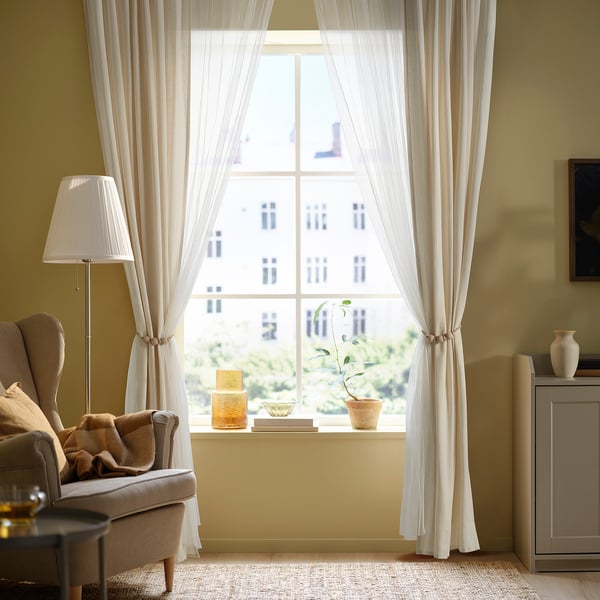 Cosy living room with TRUBBRYUM tie-backs, chair, lamp, and table decor. Curtains extend floor to ceiling.