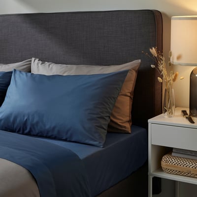 Cosy bedroom with grey bed, blue sheets, brown pillow. Bedside table with lamp, vase of dried flowers, and cushions.