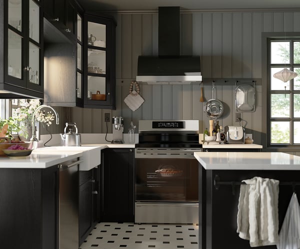 Modern kitchen with black cabinets, stainless appliances, and chequered floor. White countertops with potted plant and fruits.