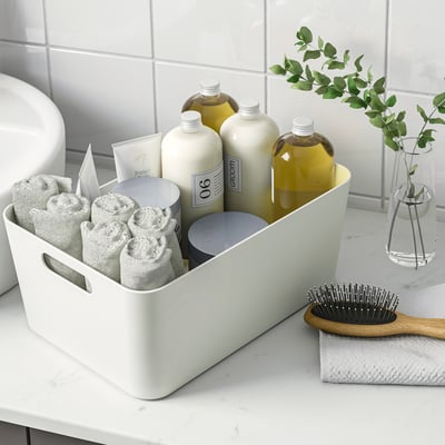 White organiser box on a bathroom counter storing neatly folded towels, shampoo bottles, and a face cream, with handles for easy portability.