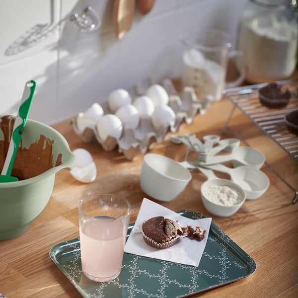 Kitchen counter with chocolate batter, eggs, measuring cups, bitten muffin & pink drink.