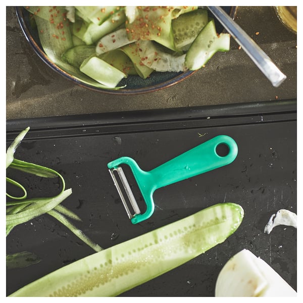 A bright green vegetable peeler rests on a black countertop next to peeled cucumbers and their skin.