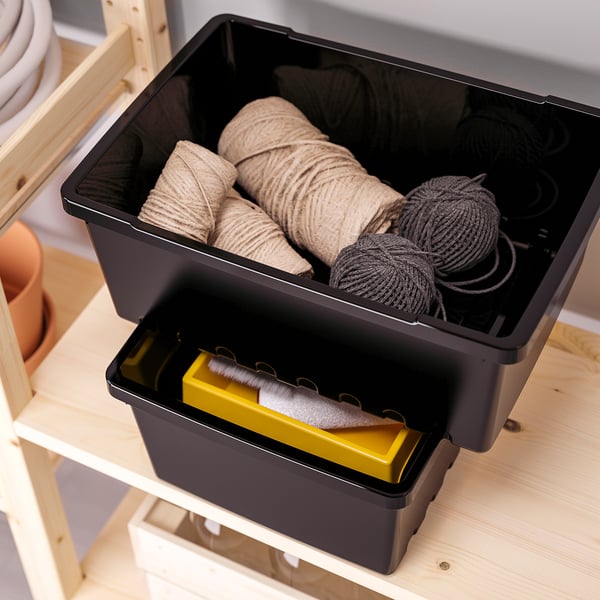 Black storage box with three compartments, holding yarn and knitting tools, placed on a wooden shelf.