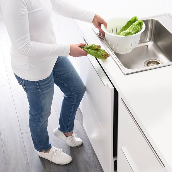 Person cutting lettuce on kitchen counter with sink and colander beside them.