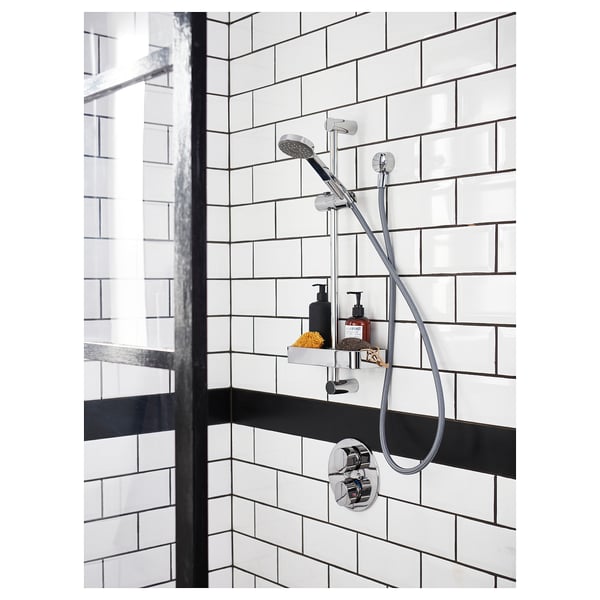 Modern shower setup with chrome VALLAMOSSE rail and handheld shower. White tiled walls, black accents, and additional shower necessities on shelf.