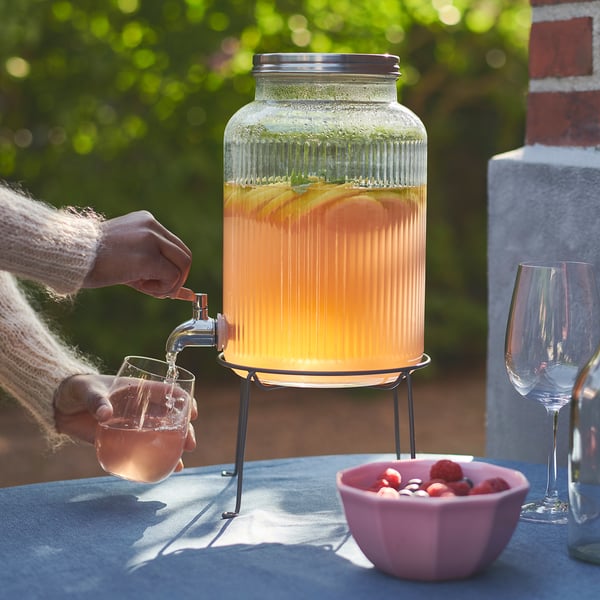 Person pours orange drink from clear VARDAGEN glass dispenser onto a glass. Bowl of fruit beside. Outdoor setting.