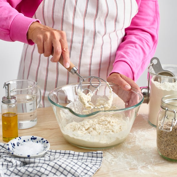 Person using danish dough whisk in glass bowl on floured surface, with ingredients nearby.