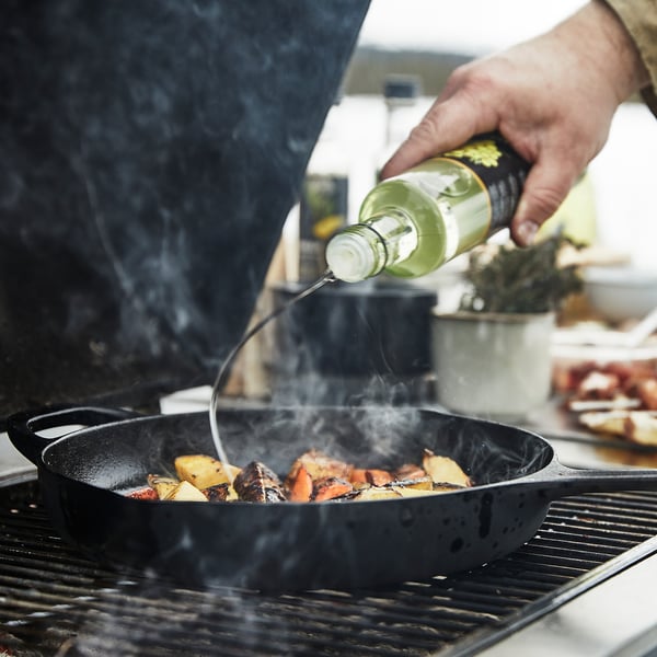 Person is drizzling oil from a green VARDAGEN bottle onto food in a black cast iron skillet on a grill. Smoke is rising from the pan.