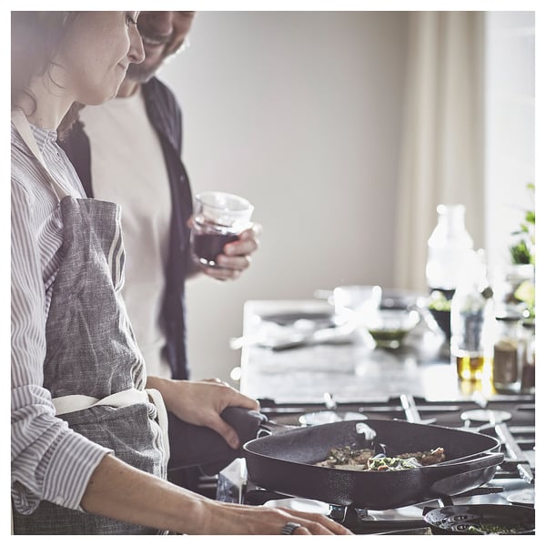A person cooks in a kitchen using a VARDAGEN cast iron black pan on a stove, holding a drink.