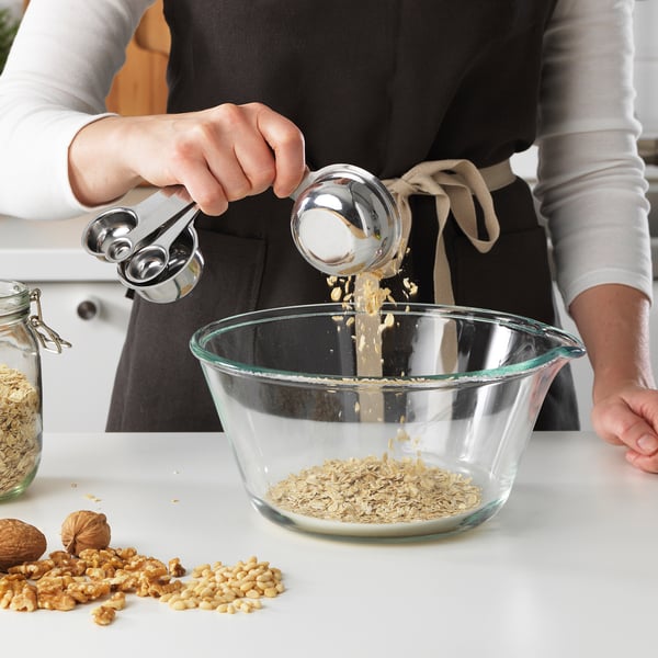 Person in apron pours ingredient into mixing bowl with measuring cups, surrounded by walnuts and oats.