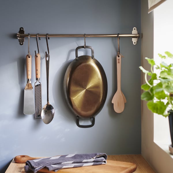 Blue wall holds rack with spatula, grater, ladle, pan, spoon. Chequered cloth and plant below.